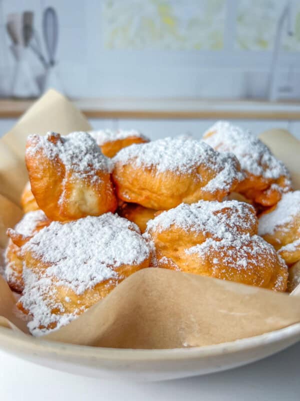 A bowl lined with parchment paper filled with golden, fluffy beignets and Mekitsi, generously dusted with powdered sugar, set on a white surface with a blurred kitchen background.