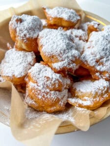 A plate lined with parchment paper holds a pile of golden brown Mekitsi, generously dusted with powdered sugar.