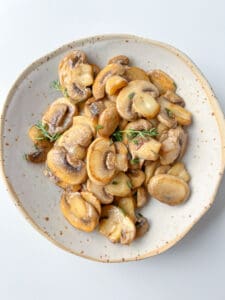A speckled ceramic plate filled with sautéed sliced mushrooms, showing How To Cook Mushrooms to perfection, garnished with fresh herbs and set on a white background.