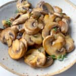 A speckled ceramic plate filled with sautéed sliced mushrooms garnished with small sprigs of fresh thyme, this image perfectly captures how to cook mushrooms for a simple yet elegant presentation, set on a light background.