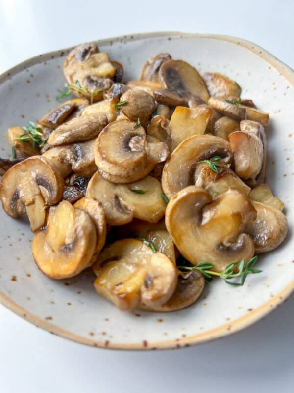 A speckled ceramic plate filled with sautéed sliced mushrooms garnished with small sprigs of fresh thyme, this image perfectly captures how to cook mushrooms for a simple yet elegant presentation, set on a light background.