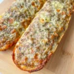 Two pieces of garlic bread topped with melted cheese and herbs, reminiscent of Bulgarian Meat Sandwiches (Prinzesi), rest on a wooden cutting board. The bread appears golden and crisp around the edges.
