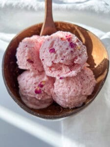 A coconut bowl filled with four scoops of light pink Cottage Cheese Ice Cream, garnished with small edible flower petals, on a white surface with sunlight casting soft shadows.