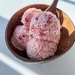 A wooden bowl filled with three scoops of pink cottage cheese ice cream, garnished with small dried flower petals, and a wooden spoon resting inside the bowl.