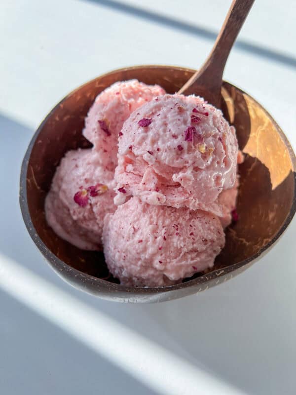 A wooden bowl filled with three scoops of pink cottage cheese ice cream, garnished with small dried flower petals, and a wooden spoon resting inside the bowl.