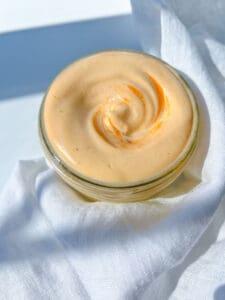 A small glass jar filled with creamy, light orange Homemade Spicy Mayo sits on a white cloth in bright, natural light.