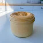 A small glass jar filled with creamy, orange-colored Homemade Spicy Mayo sits on a white countertop in a brightly lit kitchen.