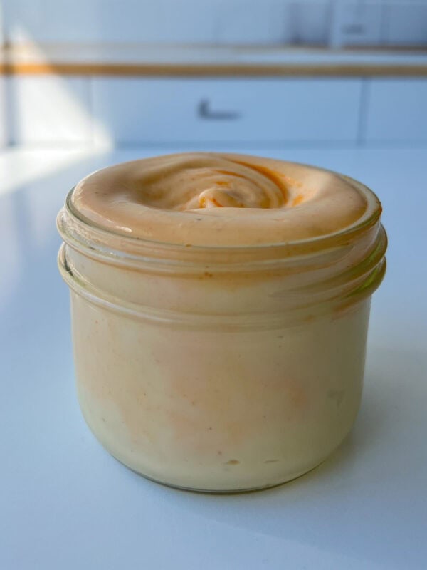 A small glass jar filled with creamy, orange-colored Homemade Spicy Mayo sits on a white countertop in a brightly lit kitchen.