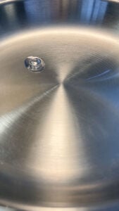A single drop of oil sits on the shiny surface of a stainless steel frying pan, reflecting light in a circular pattern—a simple step in discovering how to make stainless steel non stick.