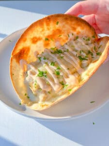 A hand holds a crispy taco shell filled with savory tuna melt, melted cheese, creamy white sauce, and chopped parsley, served on a white plate in bright sunlight.