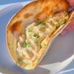 A hand holds a crispy taco shell filled with creamy, saucy mashed potatoes and hints of tuna melt flavors, topped with chopped parsley, on a white plate.