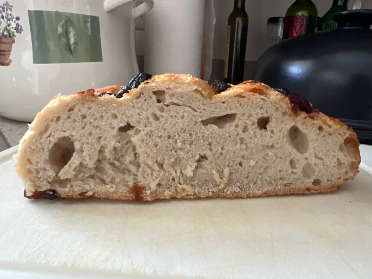 A sliced loaf of No Knead Bread with a crusty top and an airy, open crumb interior sits on a white cutting board in a kitchen.