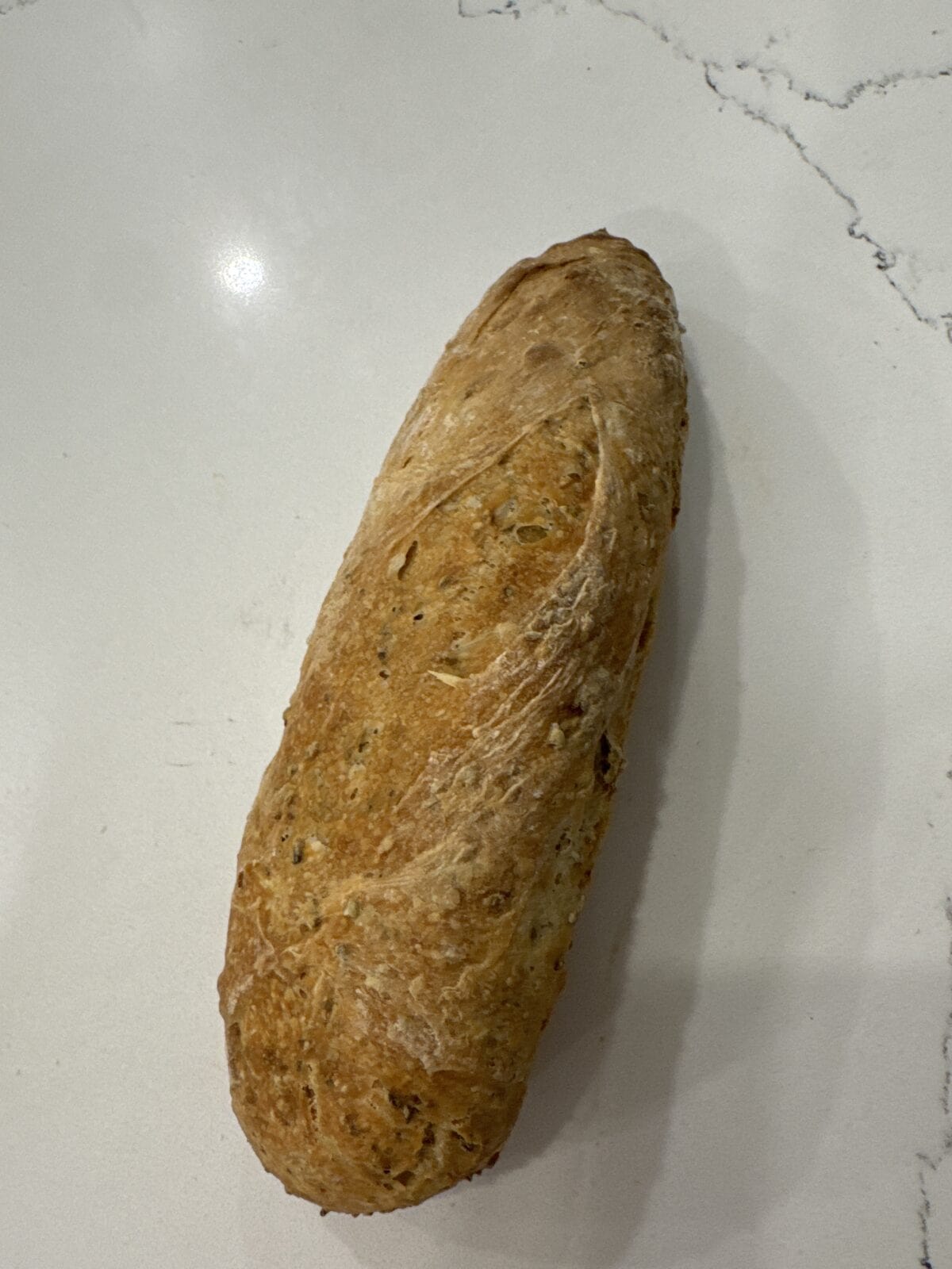 A loaf of rustic, crusty No Knead Bread with a golden-brown exterior rests on a light-colored, marbled countertop.