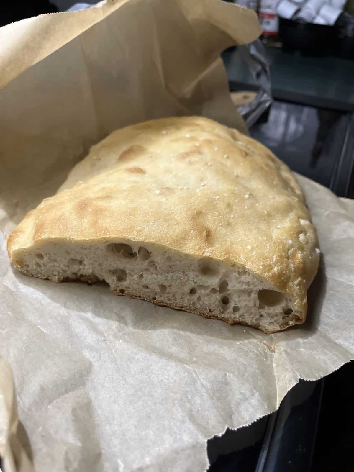 A half loaf of golden-brown, no knead focaccia bread sits on crinkled parchment paper, revealing its airy, soft interior and slightly crisp crust.