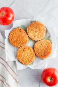 Four fried green tomato slices on a paper towel atop a marble surface, with two whole tomatoes nearby. A striped cloth is partially visible on the left side.