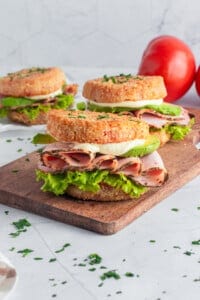 A sandwich on a wooden board features crispy breaded buns filled with sliced ham, lettuce, avocado, and cheese. In the background, there are additional sandwiches and fresh tomatoes. Chopped herbs are sprinkled on top.