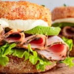 A sandwich featuring crispy, round fried bread, layered with slices of ham, bright green lettuce, and avocado, topped with creamy white sauce. The sandwich is set on a wooden board, with another similar sandwich blurred in the background.