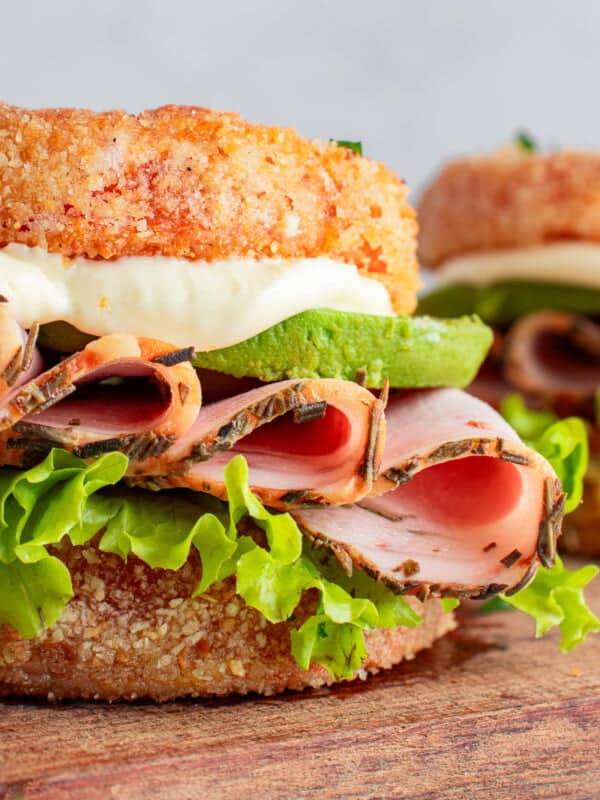 A sandwich featuring crispy, round fried bread, layered with slices of ham, bright green lettuce, and avocado, topped with creamy white sauce. The sandwich is set on a wooden board, with another similar sandwich blurred in the background.
