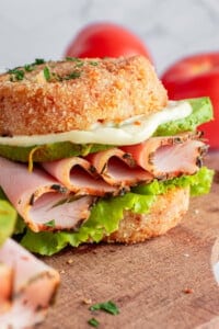 Close-up of a sandwich with lettuce, sliced turkey, avocado, and mayonnaise, served between two pieces of fried bread. In the background, there are blurred tomatoes on a wooden surface.