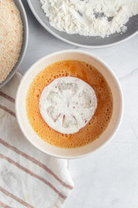 A slice of onion coated in flour sits in a bowl of beaten eggs. Nearby are plates with flour and breadcrumbs. A striped cloth is partially visible on the side. The setup suggests preparation for breading.