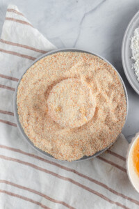 A round gray plate filled with beige breadcrumbs sits on a striped white and brown cloth. Nearby, part of a bowl with a beaten egg and another with flour are visible, suggesting preparation for breading food.