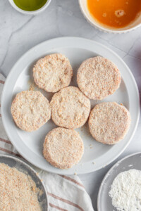 A white plate with six breaded patties, surrounded by bowls containing breadcrumbs, beaten eggs, flour, and olive oil. The scene is set on a light gray marble surface with a striped cloth.