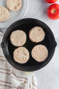 A cast iron skillet with four breaded tomato slices frying is placed on a marble surface. Two fresh tomatoes and a striped dish towel are next to the pan, with additional breaded slices on a nearby plate.
