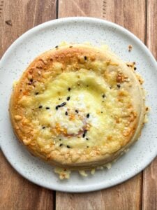 A round, baked bread topped with melted cheese and sprinkled with black and white sesame seeds, served on a white speckled plate over a wooden surface.