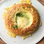 A round, sesame seed-coated bread with a melted cheese center, garnished with fresh green herbs, sits on a white speckled plate atop a wooden surface.