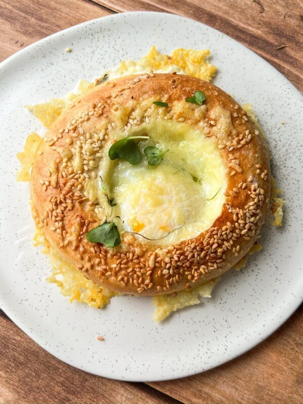 A round, sesame seed-coated bread with a melted cheese center, garnished with fresh green herbs, sits on a white speckled plate atop a wooden surface.