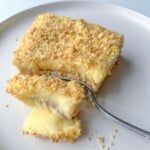 A square piece of creamy, layered dessert with a crumbly topping sits on a white plate. A fork holds a bite-sized piece, revealing a smooth yellow custard filling inside.
