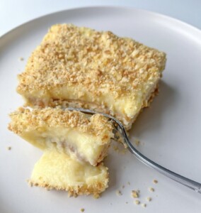 A square piece of creamy, layered dessert with a crumbly topping sits on a white plate. A fork holds a bite-sized piece, revealing a smooth yellow custard filling inside.