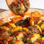 A wooden spoon holds a seasoned meatball from a dish with sliced potatoes, herbs, and bell peppers, all baked in a tomato sauce. The ingredients appear browned and garnished with fresh parsley.