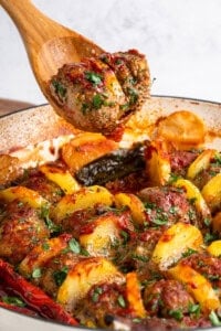 A wooden spoon holds a seasoned meatball from a dish with sliced potatoes, herbs, and bell peppers, all baked in a tomato sauce. The ingredients appear browned and garnished with fresh parsley.