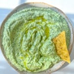 A bowl of creamy Green Goddess Dip Recipe, glistening with olive oil on top and a single tortilla chip dipped in. Bright, natural lighting highlights the herbed texture for an inviting snack.
