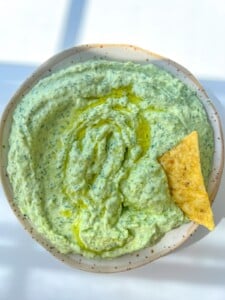 A bowl of creamy Green Goddess Dip Recipe, glistening with olive oil on top and a single tortilla chip dipped in. Bright, natural lighting highlights the herbed texture for an inviting snack.