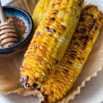 Three grilled ears of corn on the cob rest on parchment paper in a dish, with a small bowl of hot honey butter and a honey dipper beside them. The corn has a golden, lightly charred appearance—perfect Hot Honey Butter Corn.