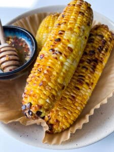 Three grilled ears of corn on the cob rest on parchment paper in a dish, with a small bowl of hot honey butter and a honey dipper beside them. The corn has a golden, lightly charred appearance—perfect Hot Honey Butter Corn.
