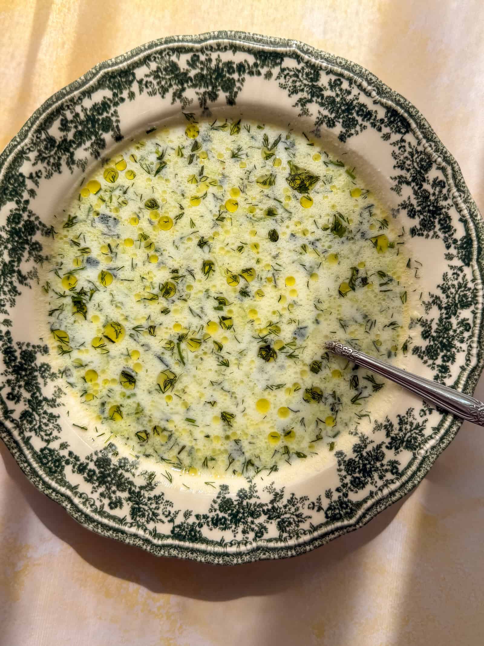 A bowl of creamy Cold Cucumber Soup with chopped green herbs and drops of oil on top, served in a decorative bowl with a green floral pattern and a metal spoon on a light-colored surface.