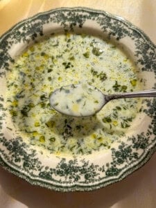 A close-up of a bowl of creamy Cold Cucumber Soup with herbs and oil, served on a vintage white plate with a green floral rim. A spoonful of the chilled soup is held above the bowl, catching the sunlight.