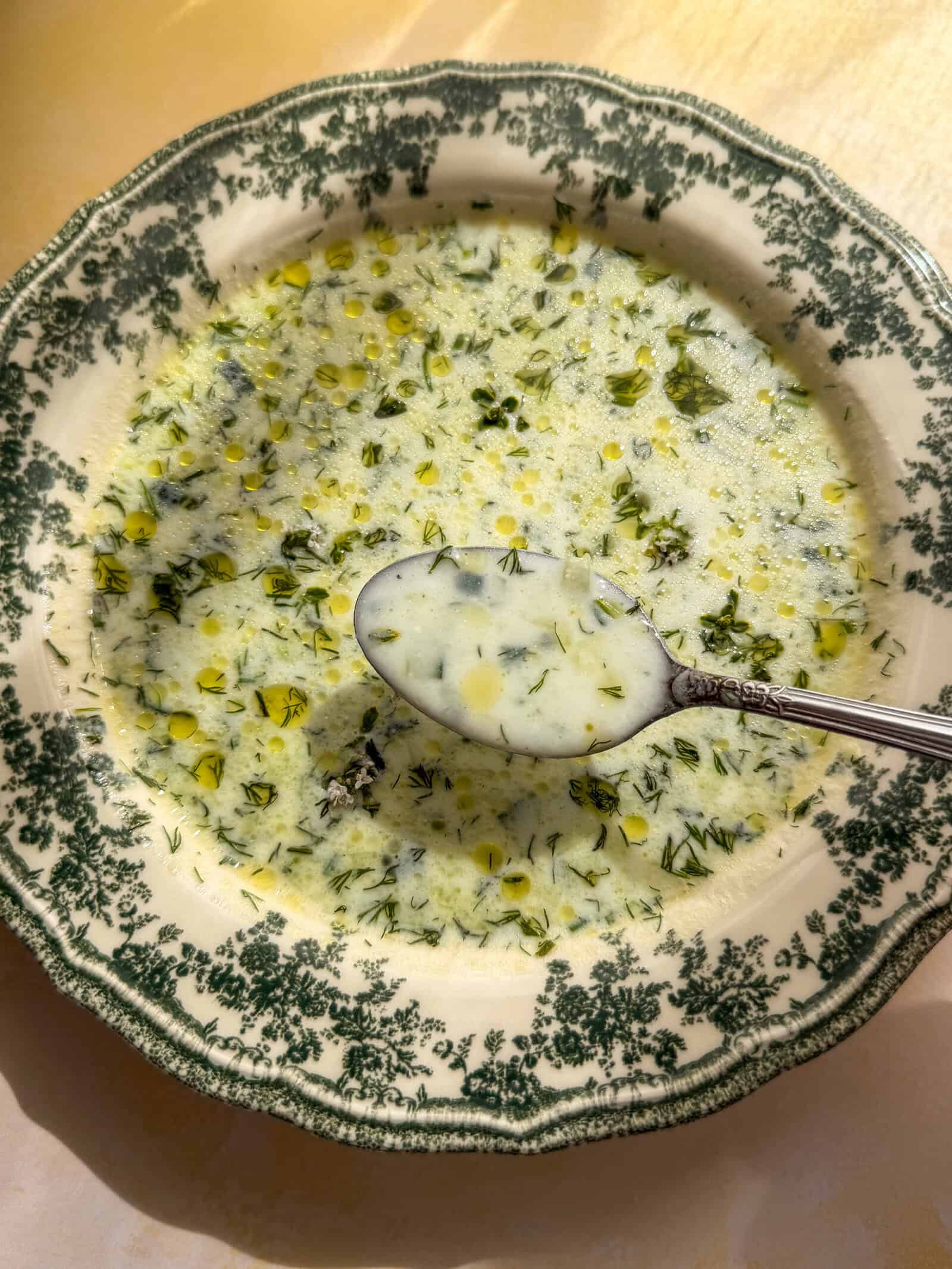 A bowl of creamy Cold Cucumber Soup with green herbs and yellow oil droplets, served in a decorative green and white bowl. A spoon rests in the soup, holding some liquid and herbs as sunlight shines on part of the bowl.
