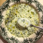 A decorative bowl filled with creamy, herb-speckled Cold Cucumber Soup, garnished with oil drops and fresh dill, with a spoon lifting some of the soup. Warm light highlights the texture and refreshing details.