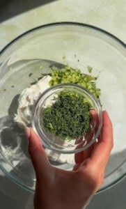 A hand holds a small bowl of chopped fresh dill over a glass mixing bowl containing mayonnaise and chopped cucumbers, the perfect base for a refreshing Cold Cucumber Soup.
