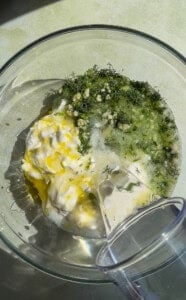 A glass bowl contains yogurt, grated cucumber, minced garlic, chopped dill, olive oil, and vinegar, with a measuring cup pouring in liquid—classic ingredients for tzatziki sauce or a refreshing Cold Cucumber Soup—on a light surface.