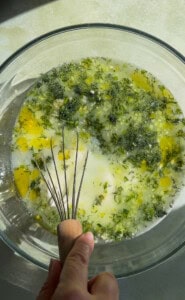 A hand holding a whisk in a glass bowl filled with oil, chopped herbs, and liquid ingredients—perfect for preparing Cold Cucumber Soup—with sunlight casting gentle shadows across the surface.