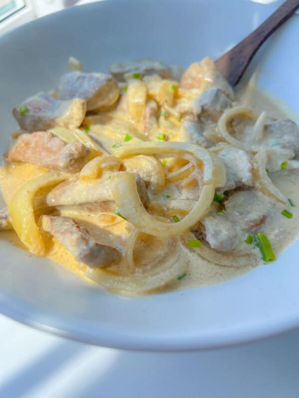 Bowl of creamy stew featuring a classic Liver and Onions Recipe, with tender meat slices, onions, and light sauce, garnished with chopped green herbs and served with a wooden spoon.