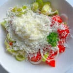 A white bowl filled with sliced tomatoes, cucumbers, onions, and peppers is topped with a generous amount of grated white cheese and parsley—classic ingredients of a traditional Shopska Salad.
