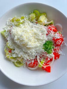A white bowl filled with sliced tomatoes, cucumbers, onions, and peppers is topped with a generous amount of grated white cheese and parsley—classic ingredients of a traditional Shopska Salad.