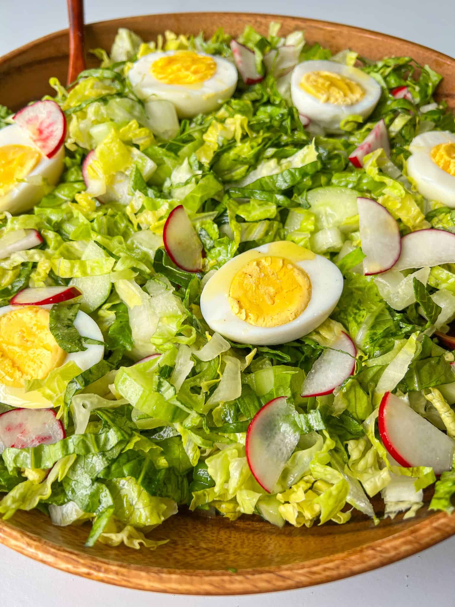A fresh salad in a wooden bowl featuring sliced hard-boiled eggs, radishes, and cucumbers on a bed of chopped lettuce.