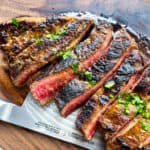 Sliced medium-rare steak with a seared crust, inspired by a classic Carne Asada recipe, garnished with chopped herbs and displayed on a wooden cutting board with a knife partially visible underneath.